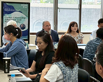 室內會議場景：中央男子手持麥克風發言，前方與會者在桌邊討論與閱讀資料；背景海報寫有「Global Citizenship Education and Sustainability，2023.5.22」，大面窗戶引入自然光。]