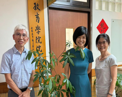 室內合照，左起：銀髮戴眼鏡男（淺藍短袖）、短髮女（青綠洋裝）、長髮紮馬尾女（淺灰上衣與褲）；背景有綠色盆栽與木質中文招牌，氣氛明亮友善。