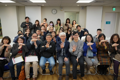 約30人於室內交流場合合照；前排坐著、後排站立，皆做水平手勢，氣氛友善合作，背景有時鐘與門，木地板與排椅可見。