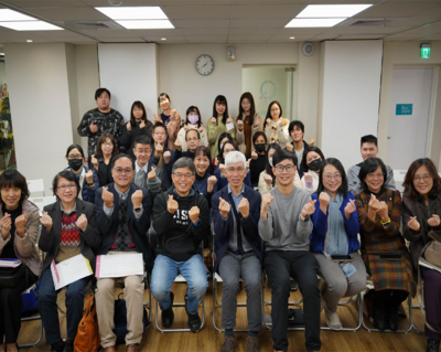 約30人於室內交流場合合照；前排坐著、後排站立，皆做水平手勢，氣氛友善合作，背景有時鐘與門，木地板與排椅可見。