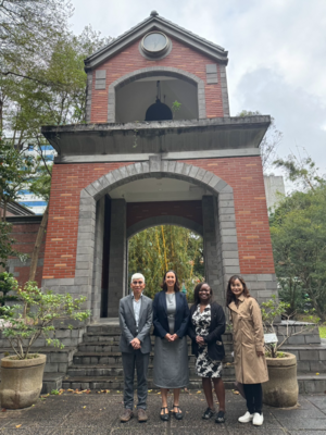 四人於紅磚拱門鐘樓前合照；左起灰色西裝男、灰裙深色外套女、黑白圖樣洋裝配黑外套女、米色大衣黑褲白鞋女。背景有時鐘、懸掛鐘、盆栽與樹木，天空多雲。